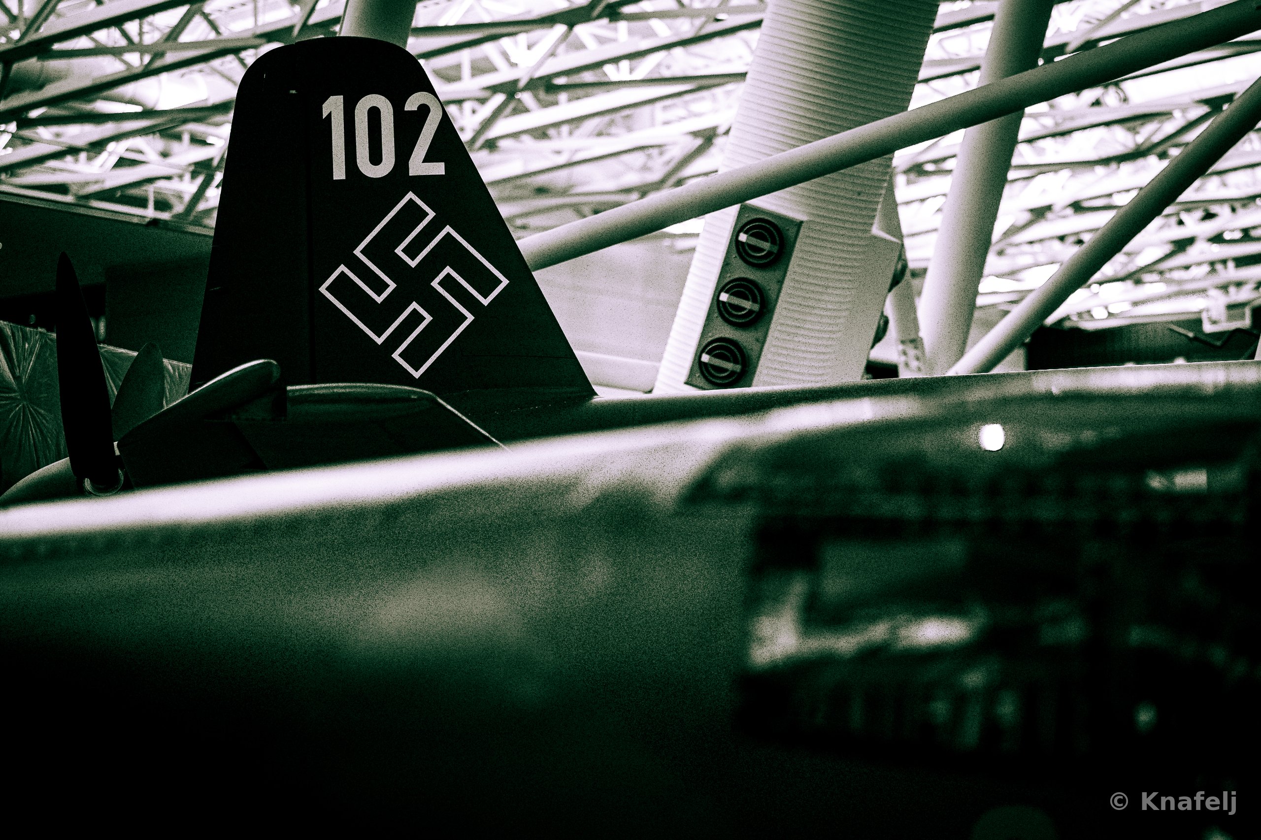 Black and white photograph of an aircraft tail fin numbered 102 with historical wartime markings, shot from below against a museum's geometric metal roof structure.