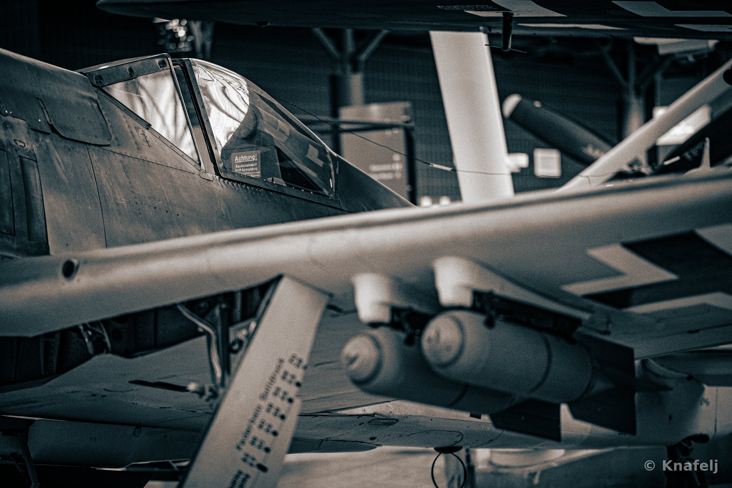 Monochrome image of a historic military aircraft cockpit with glass canopy, partially obscured by the wing and bomb-shaped pods of another plane in a dimly lit museum hangar.