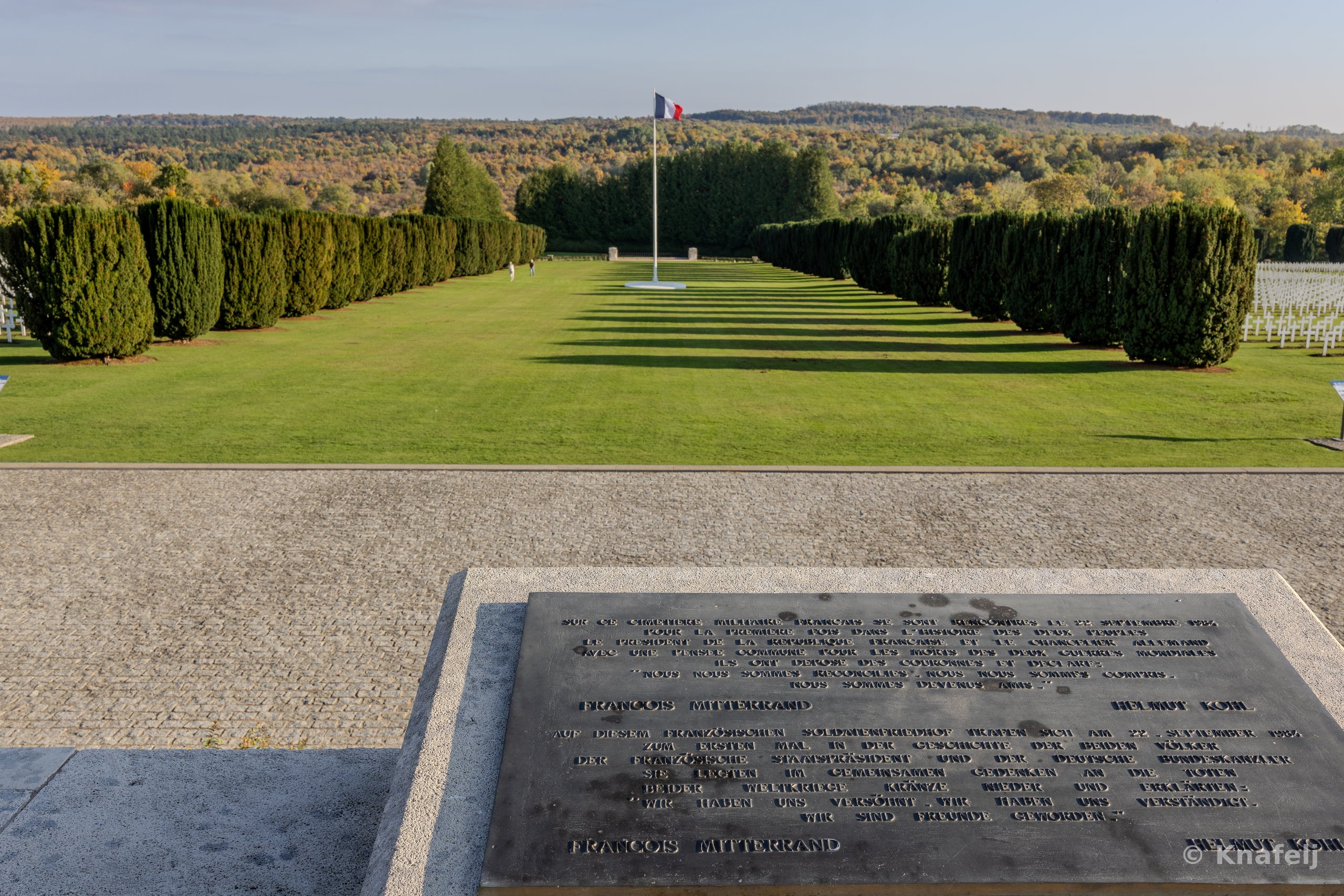 A bronze memorial plaque inscribed in French and German in the foreground, with a French flag on a tall pole, neatly trimmed conifer trees, white crosses, and a green lawn leading to forested hills in the background.