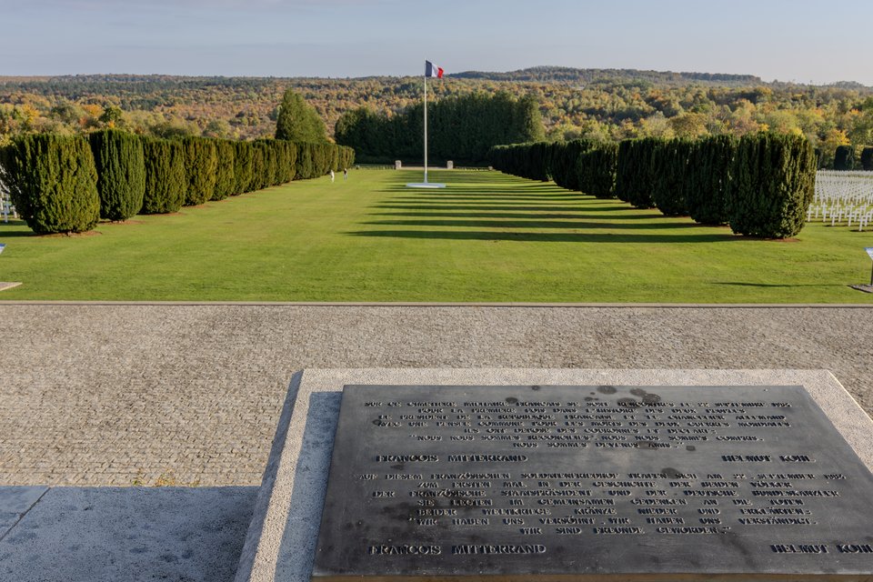 A bronze memorial plaque inscribed in French and German in the foreground, with a French flag on a tall pole, neatly trimmed conifer trees, white crosses, and a green lawn leading to forested hills in the background.