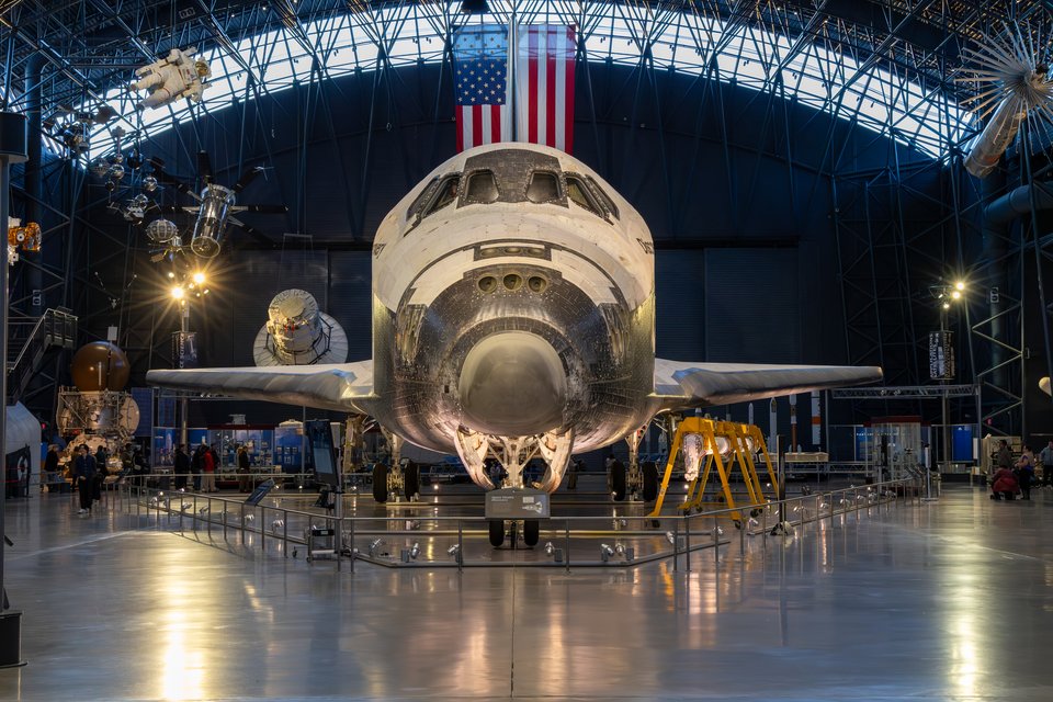 Front view of a space shuttle orbiter displayed in a large museum hangar, with an American flag hanging behind it, other space artifacts suspended overhead, and a reflective polished floor.