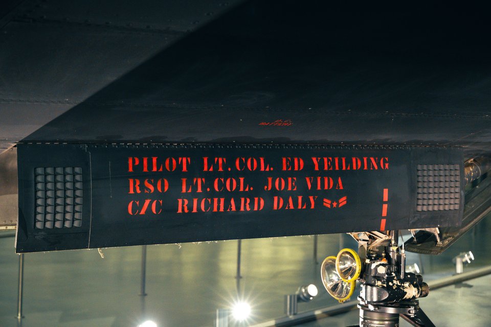 Underbelly of a dark SR-71 aircraft with red stenciled text reading PILOT LT. COL. ED YEILDING, RSO LT. COL. JOE VIDA, and C/C RICHARD DALY, with landing gear and lights visible below.