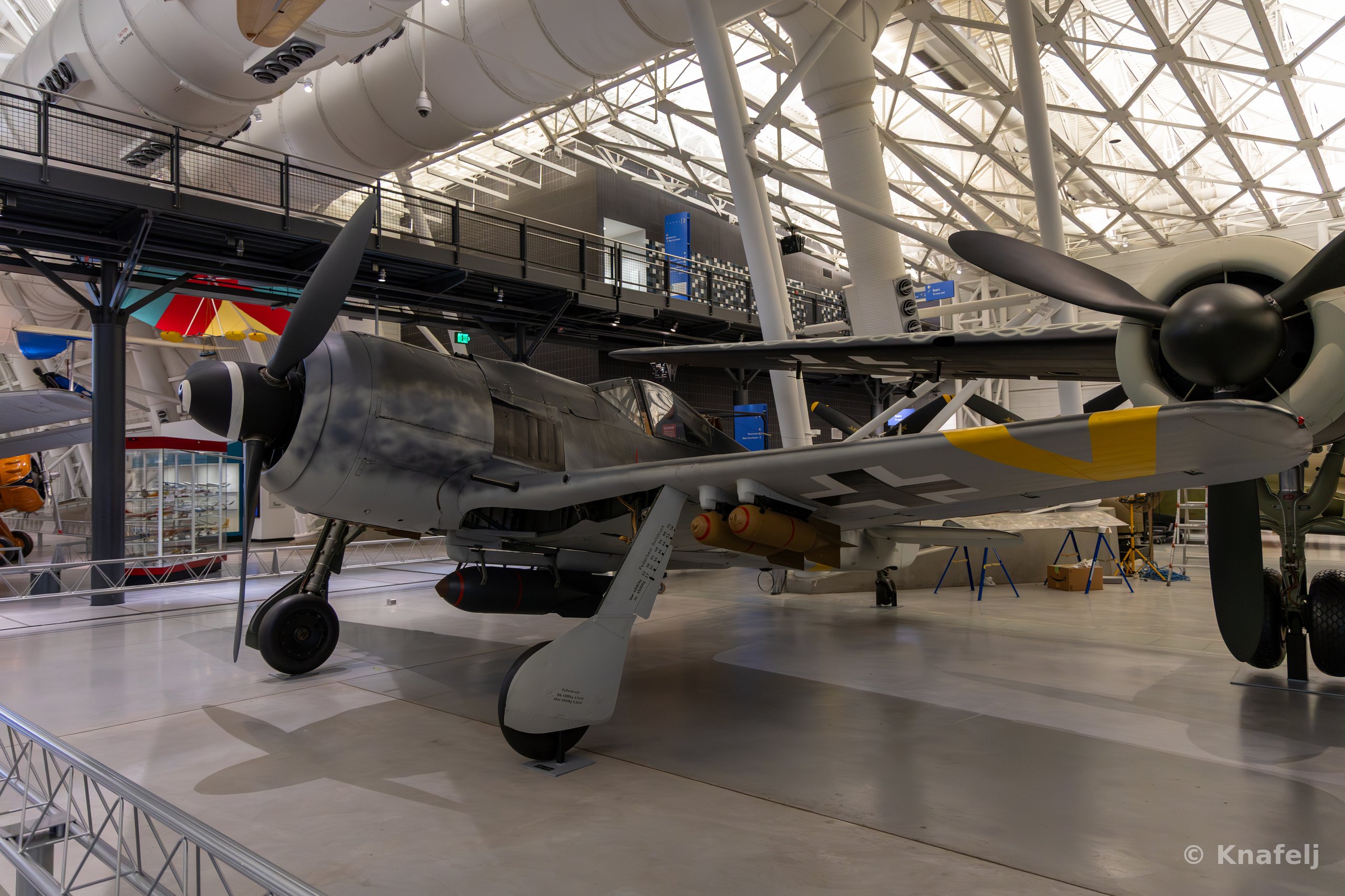 A World War II era Focke-Wulf 190 fighter plane with gray fuselage and yellow tail markings displayed indoors in a large museum hangar with a curved metal-framed glass ceiling.