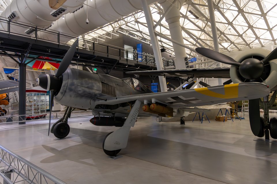 A World War II era Focke-Wulf 190 fighter plane with gray fuselage and yellow tail markings displayed indoors in a large museum hangar with a curved metal-framed glass ceiling.