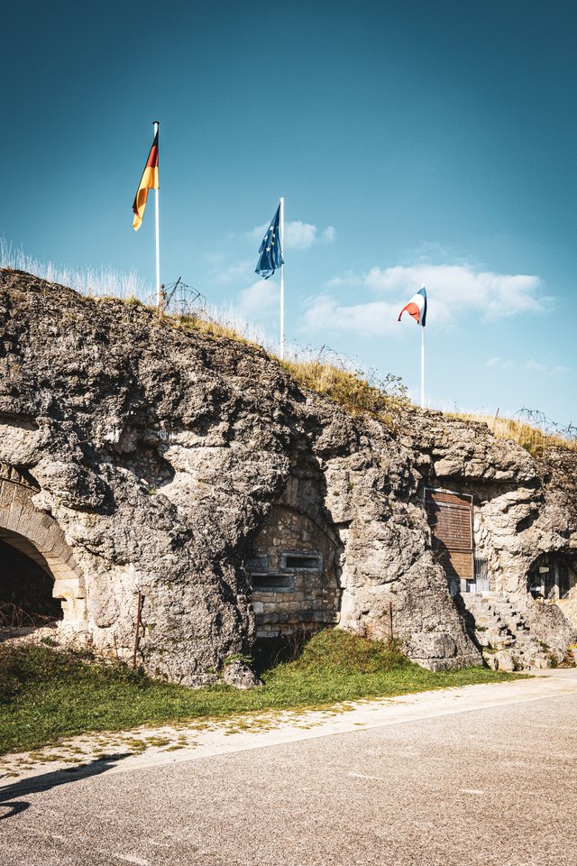 A rugged stone fortress with grass on top, beneath three flagpoles flying the German, EU, and French flags against a blue sky with light clouds.