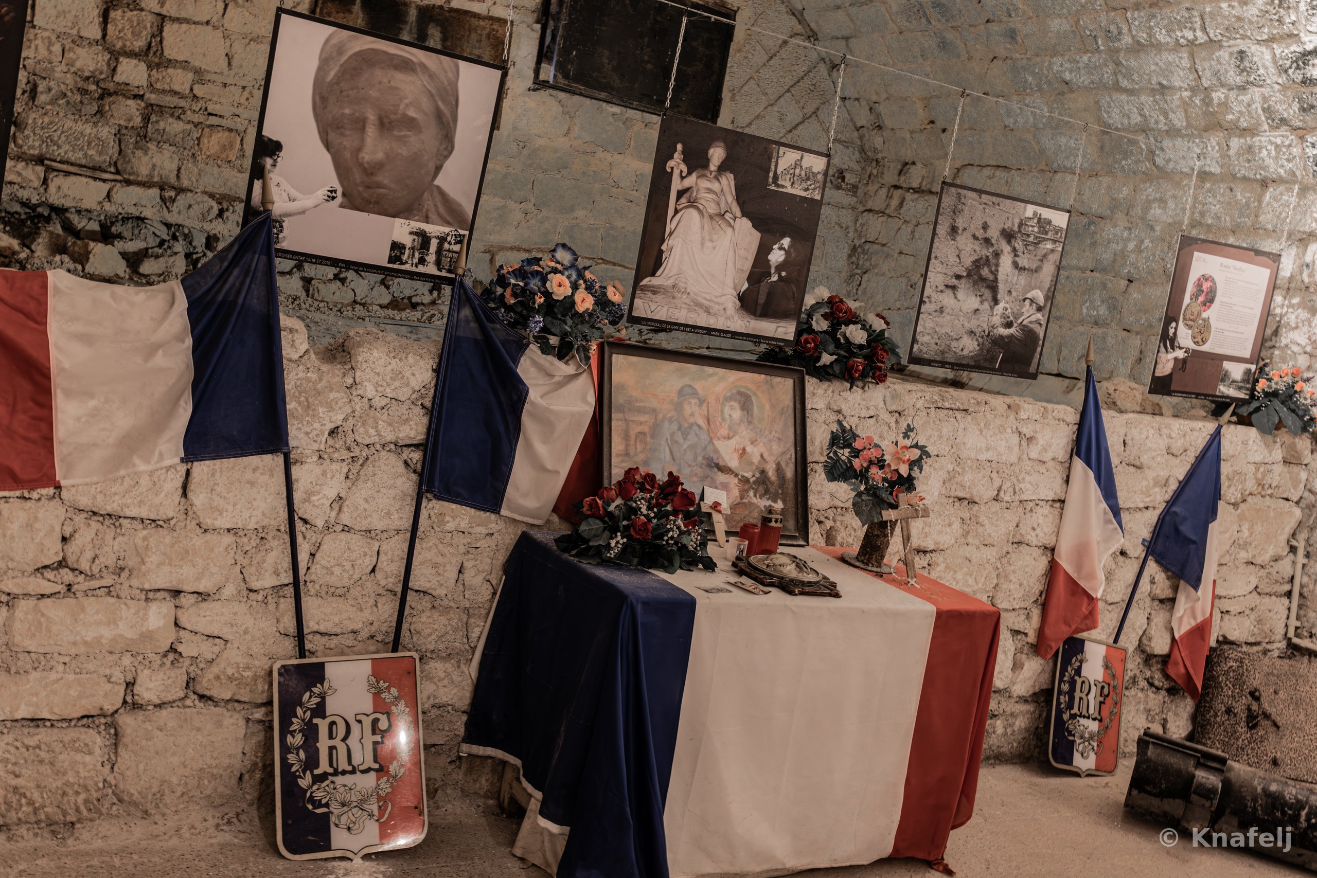 A dimly lit stone-walled room with French tricolor flags draped on a table covered with flowers, framed pictures, and RF shield emblems, surrounded by historic black-and-white photographs mounted on the walls.