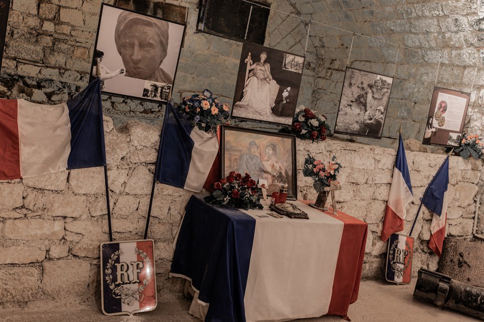 A dimly lit stone-walled room with French tricolor flags draped on a table covered with flowers, framed pictures, and RF shield emblems, surrounded by historic black-and-white photographs mounted on the walls.