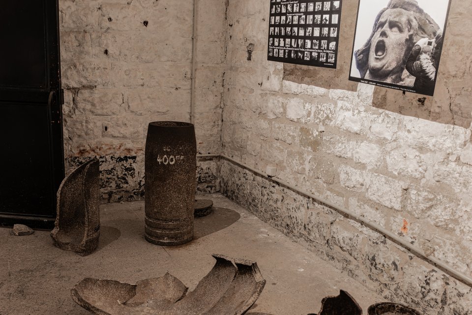 A heavy 400mm artillery shell labeled in white paint stands upright on a concrete floor surrounded by broken metal fragments, with whitewashed brick walls behind it and black-and-white war photographs mounted on the wall.