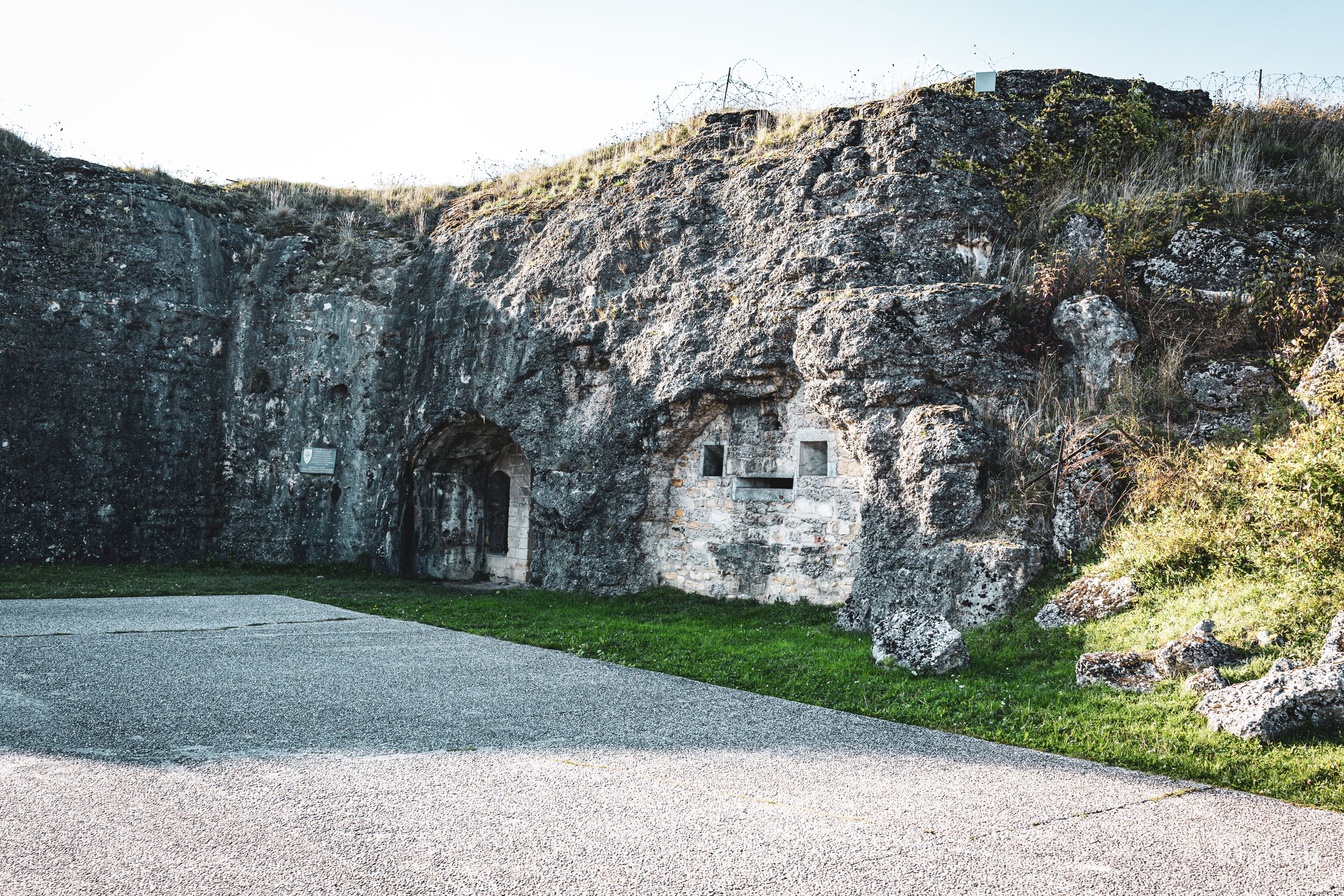 A weathered stone fortress structure built into a grassy hill, with eroded walls, a dark arched doorway, and small window openings, set above a paved courtyard and patches of green grass.