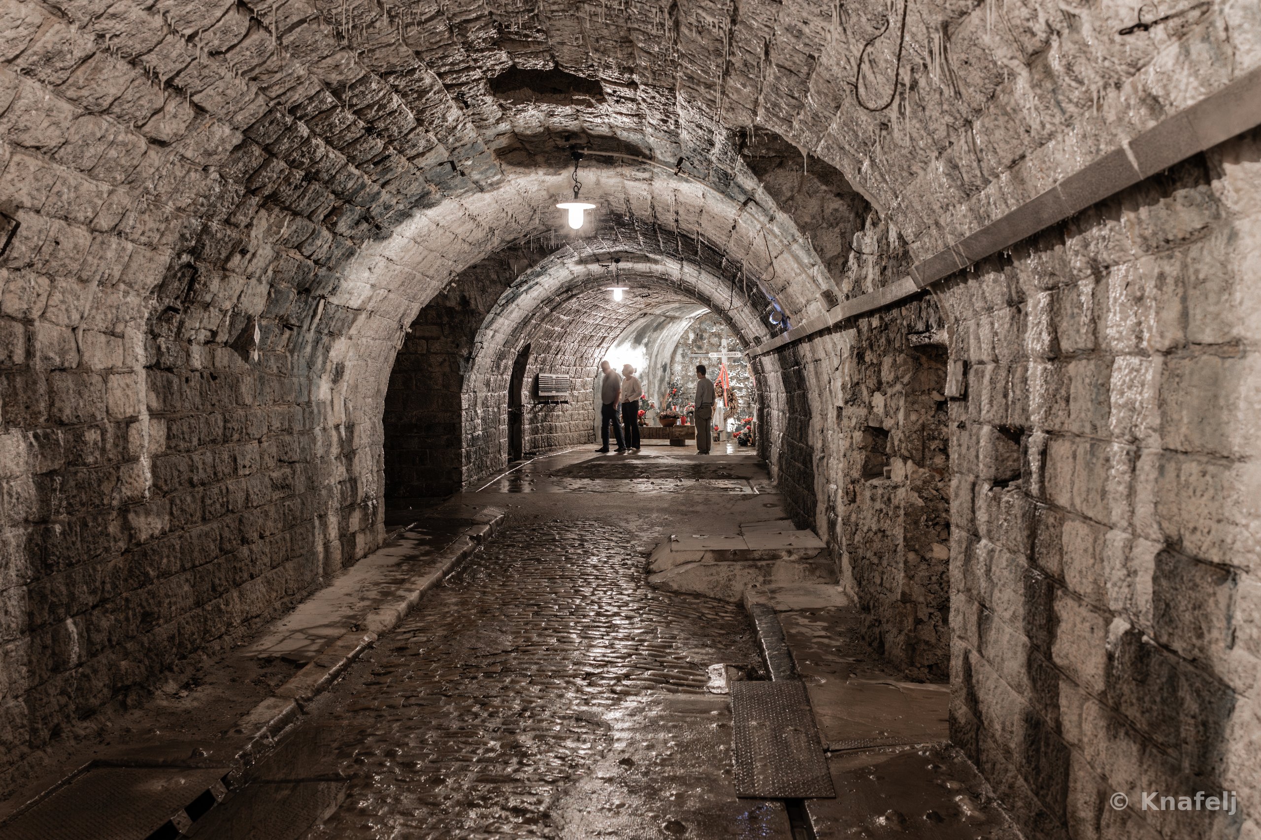 An underground stone tunnel with arched ceilings and lamps, leading toward several people standing in the distance on a wet cobblestone floor.