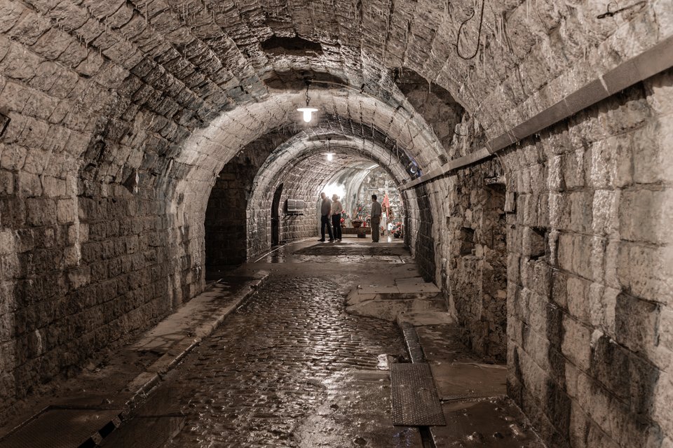 An underground stone tunnel with arched ceilings and lamps, leading toward several people standing in the distance on a wet cobblestone floor.