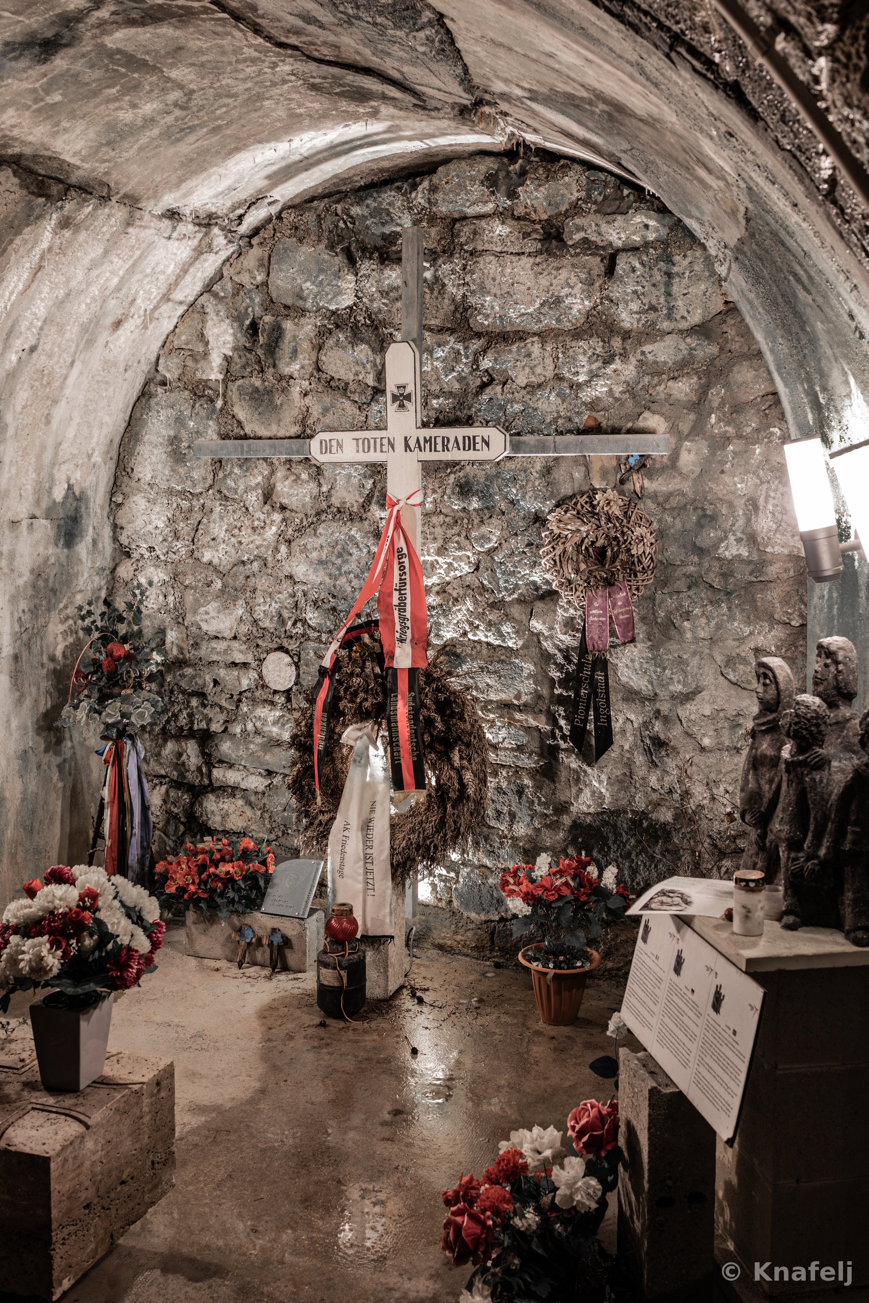 Inside a stone-walled vaulted chamber, a metal cross labeled 'Den Toten Kameraden' stands against an old brick wall, surrounded by funeral wreaths, red and black ribbons, flower arrangements, candles, and a carved stone sculpture of soldiers.
