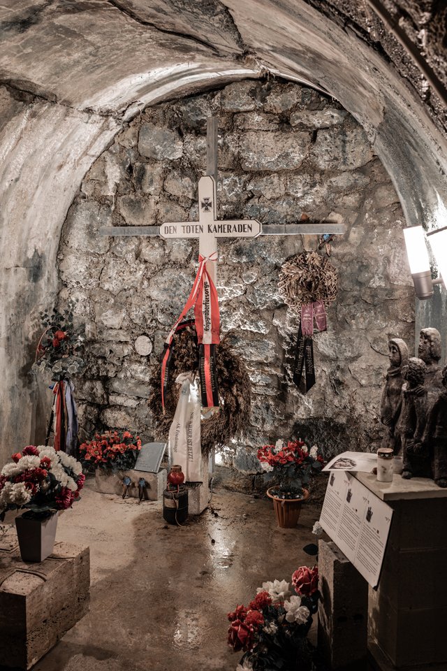 Inside a stone-walled vaulted chamber, a metal cross labeled 'Den Toten Kameraden' stands against an old brick wall, surrounded by funeral wreaths, red and black ribbons, flower arrangements, candles, and a carved stone sculpture of soldiers.