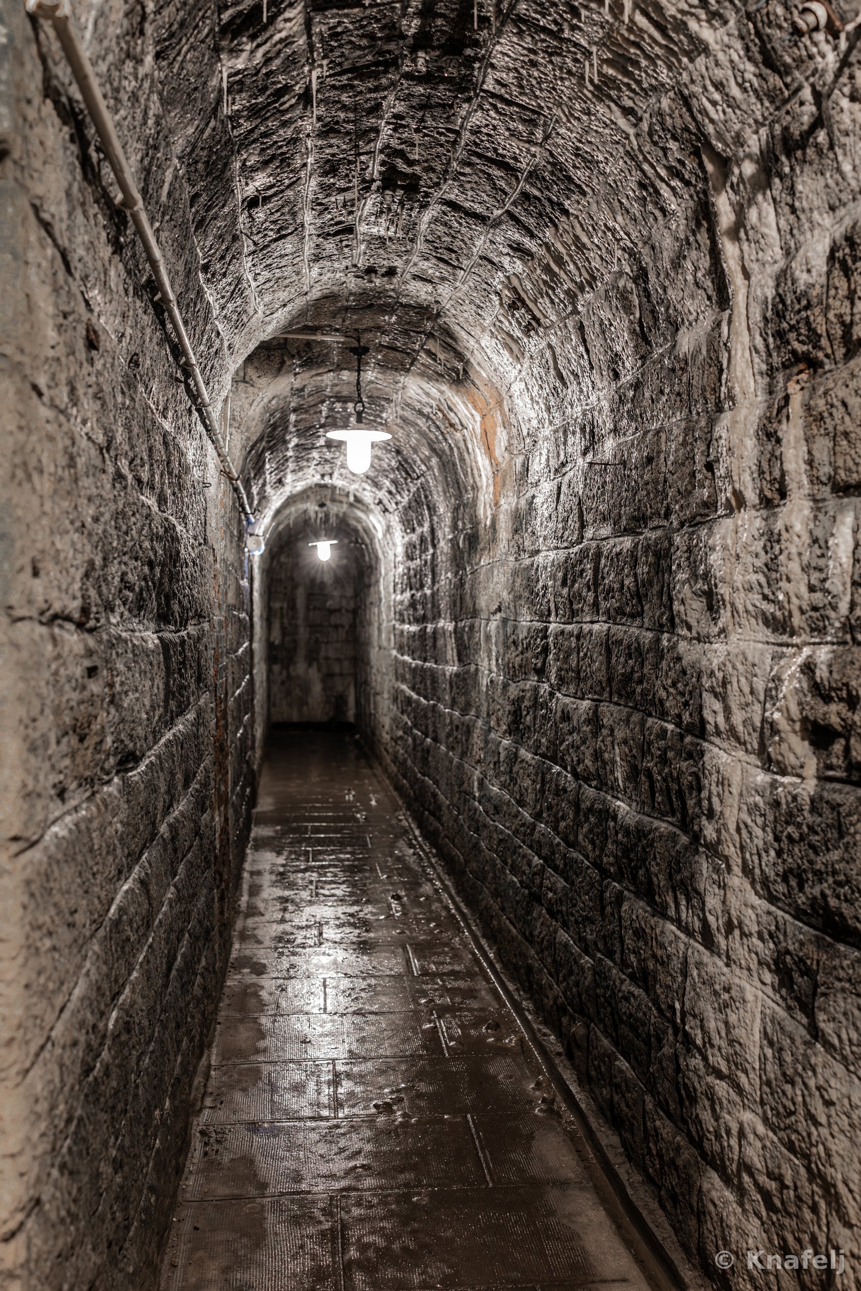 An underground arched tunnel built of weathered stone blocks, lit by two hanging ceiling lamps, with a wet reflective floor leading into the dark distance.