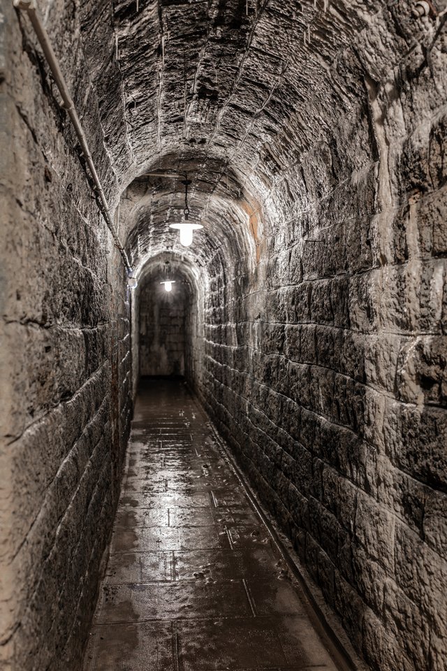 An underground arched tunnel built of weathered stone blocks, lit by two hanging ceiling lamps, with a wet reflective floor leading into the dark distance.