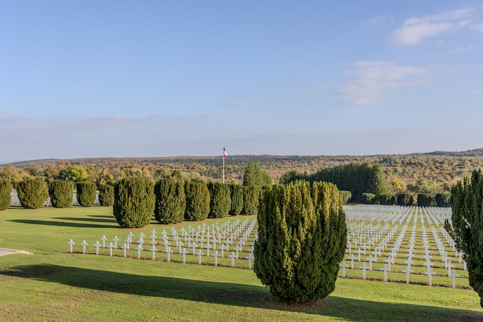 A war cemetery with many white crosses laid out in rows on green grass, separated by rounded evergreen shrubs, with a French flag and tree-covered hills under a blue sky in the background.