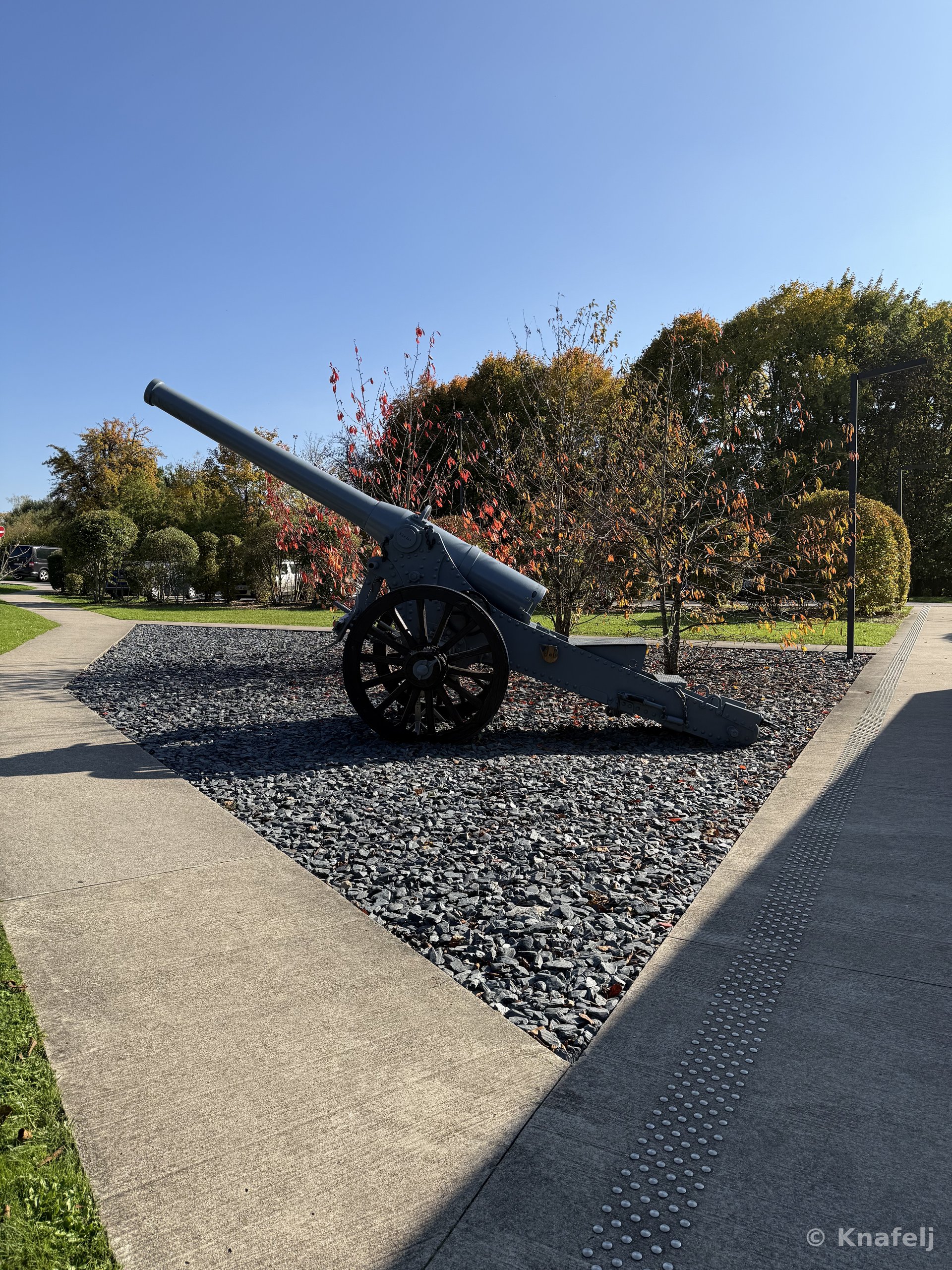 An old gray military cannon with large spoked wheels mounted on a gravel display area, surrounded by trees with orange and green autumn leaves and a paved walkway under a clear blue sky.