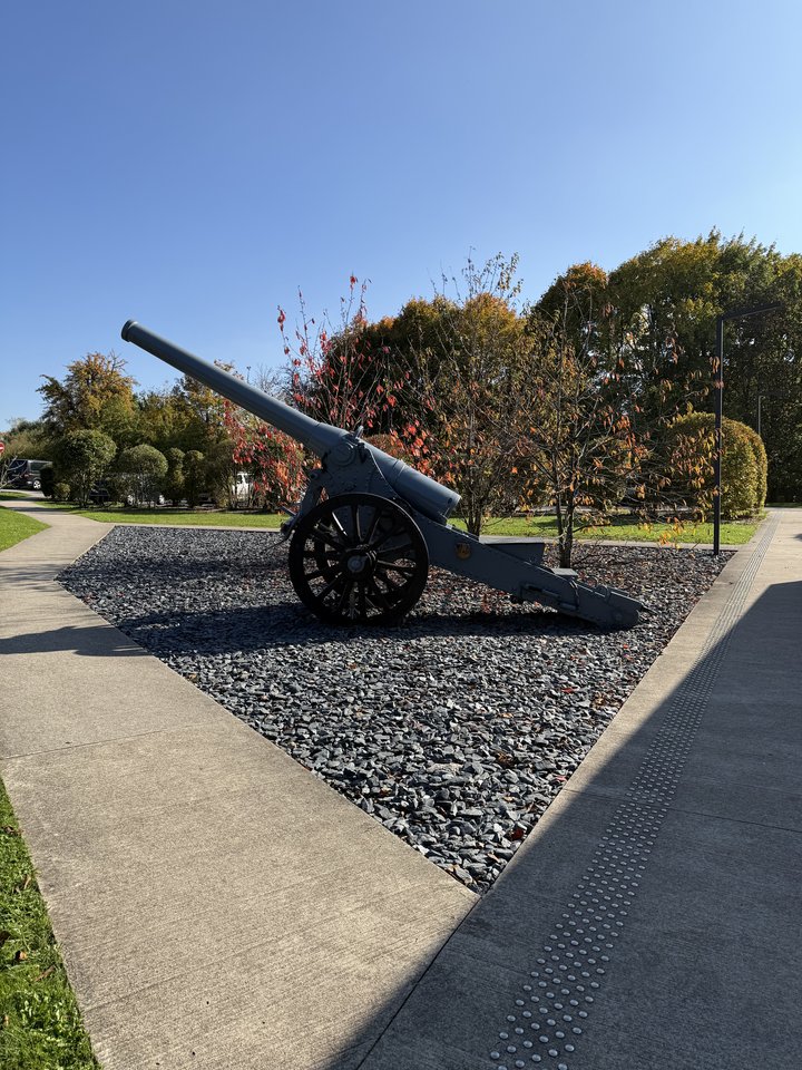 An old gray military cannon with large spoked wheels mounted on a gravel display area, surrounded by trees with orange and green autumn leaves and a paved walkway under a clear blue sky.