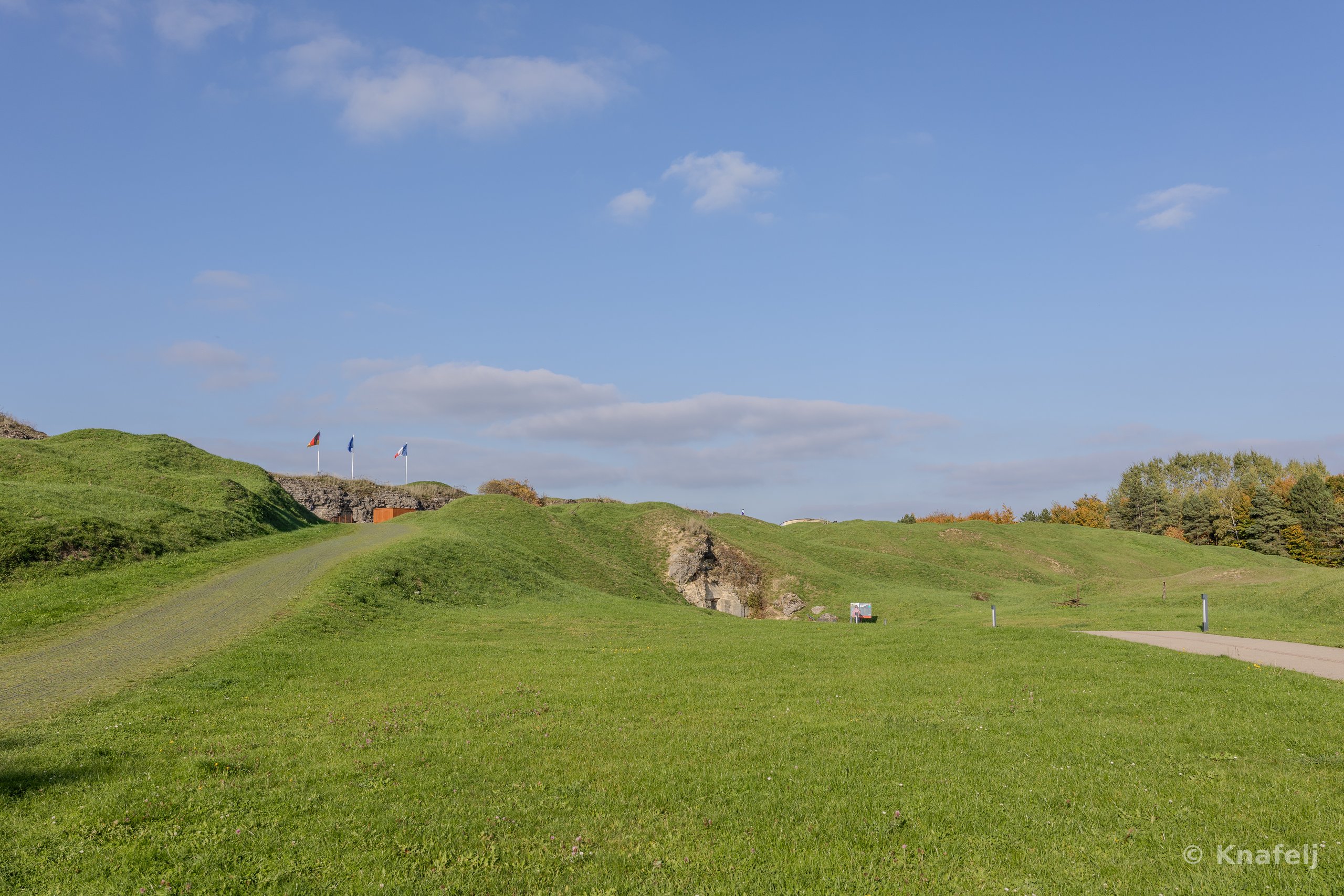 Green grassy hills under a bright blue sky with scattered clouds, with three flags flying on a distant ridge and a paved path curving through the landscape near a line of trees.