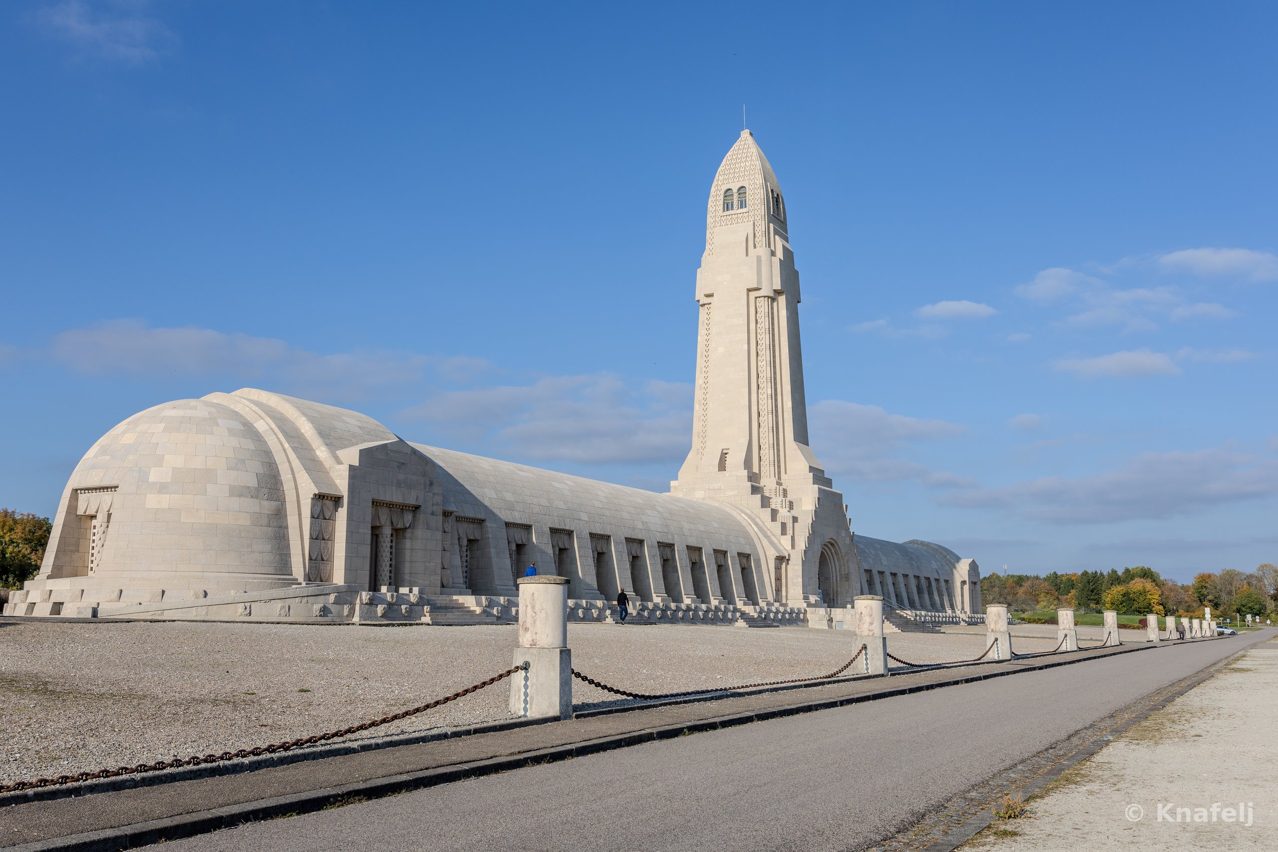 A large pale stone war memorial with a tall central tower and a long arched vaulted building, bordered by a low chain barrier and an empty path, under a clear blue sky.