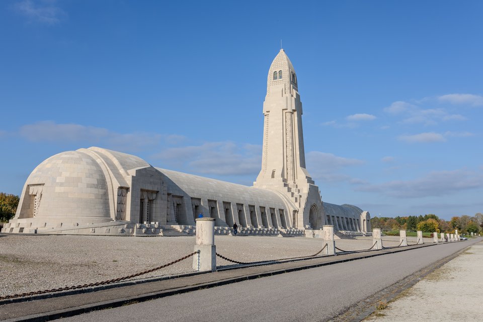 A large pale stone war memorial with a tall central tower and a long arched vaulted building, bordered by a low chain barrier and an empty path, under a clear blue sky.
