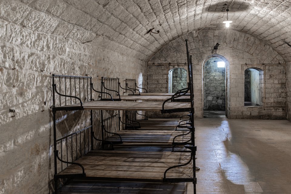 Interior of a vaulted stone room with arched ceiling containing multiple wrought-iron bunk beds with wooden slats, arched doorways and windows in the background, and a single light bulb hanging from the ceiling.