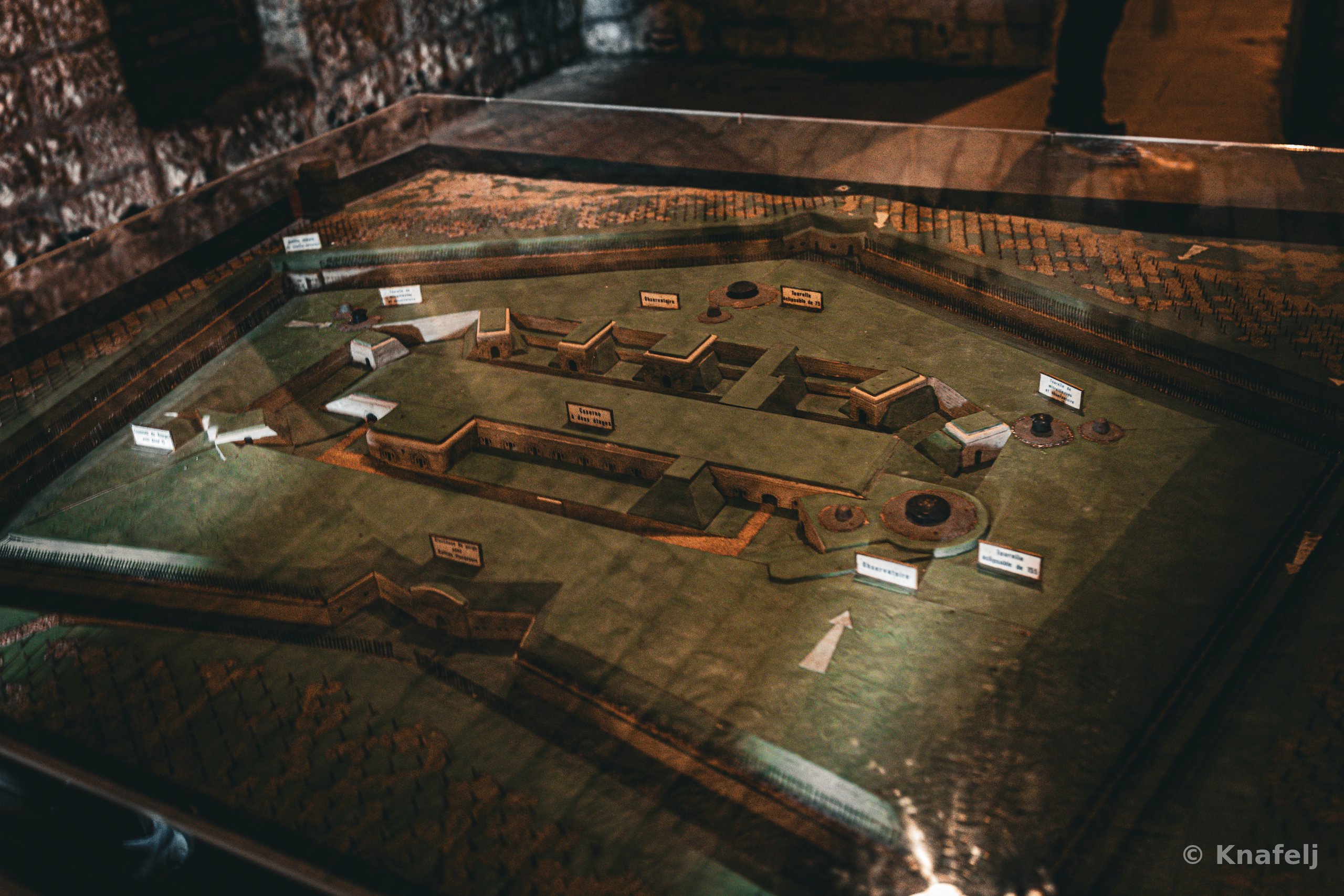 A scale model of a fortified military complex displayed under glass in a museum, showing walls, buildings, and gun emplacements with small white identification labels, illuminated by warm low light.