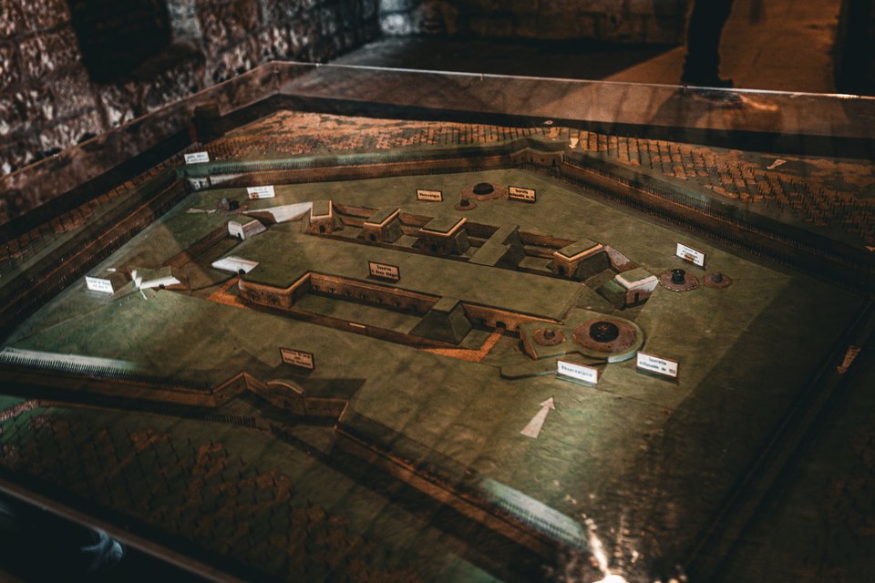 A scale model of a fortified military complex displayed under glass in a museum, showing walls, buildings, and gun emplacements with small white identification labels, illuminated by warm low light.
