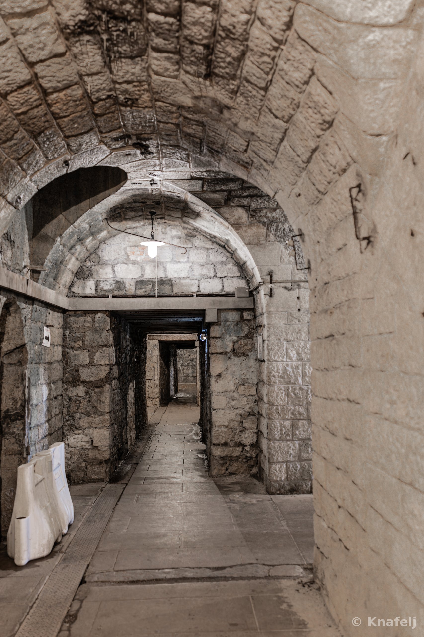 An ancient vaulted stone corridor with arched ceilings and stone block walls, leading down a flagstone path into a dimly lit distance, with a single light fixture hanging from the ceiling.