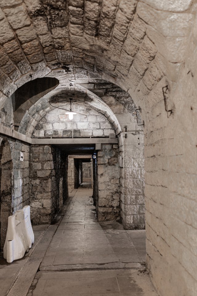 An ancient vaulted stone corridor with arched ceilings and stone block walls, leading down a flagstone path into a dimly lit distance, with a single light fixture hanging from the ceiling.