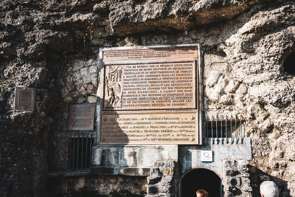 Bronze plaques with French inscriptions set into a rough stone cliff wall above a dark tunnel entrance, with two people visible at the bottom of the frame.