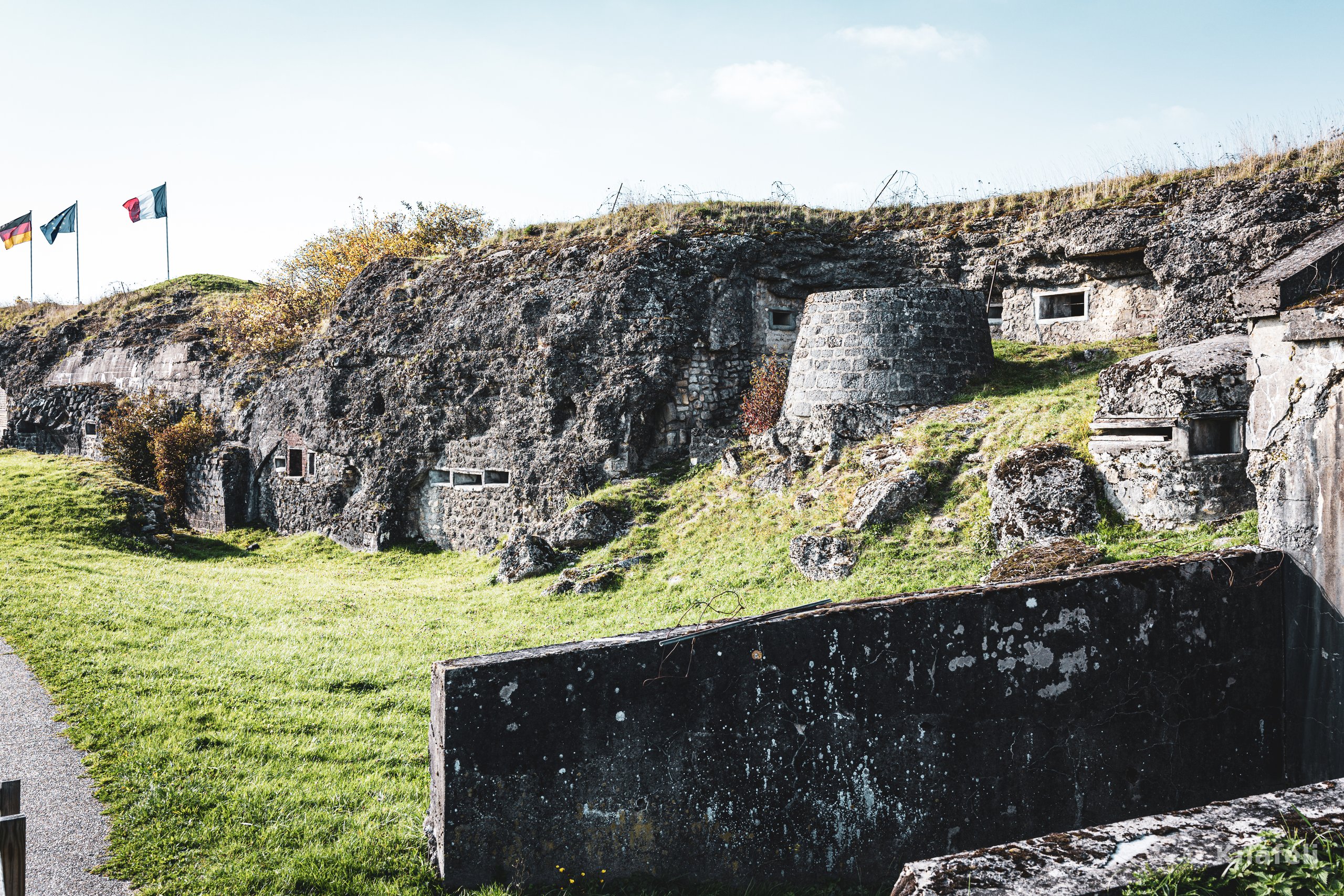 Ruins of an old stone military fort overgrown with grass and moss, with embrasures and bunkers visible. Three flags fly on poles at the upper left, and a low concrete wall sits in the foreground.