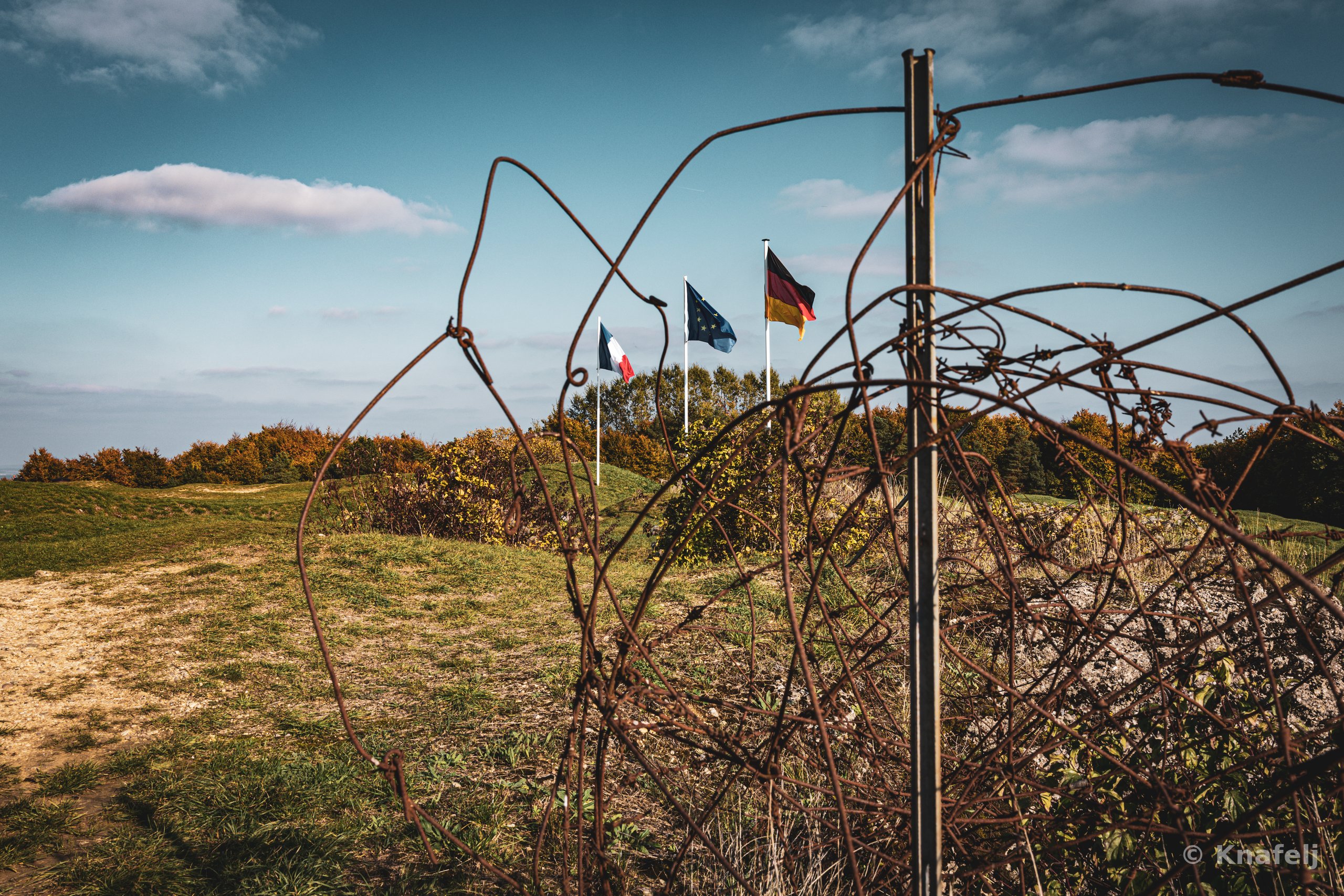 French, EU, and German flags on poles in a grassy field, viewed through twisted rusty barbed wire with a metal post in the foreground and autumn shrubs on the horizon.