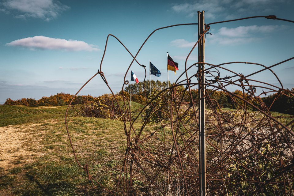French, EU, and German flags on poles in a grassy field, viewed through twisted rusty barbed wire with a metal post in the foreground and autumn shrubs on the horizon.