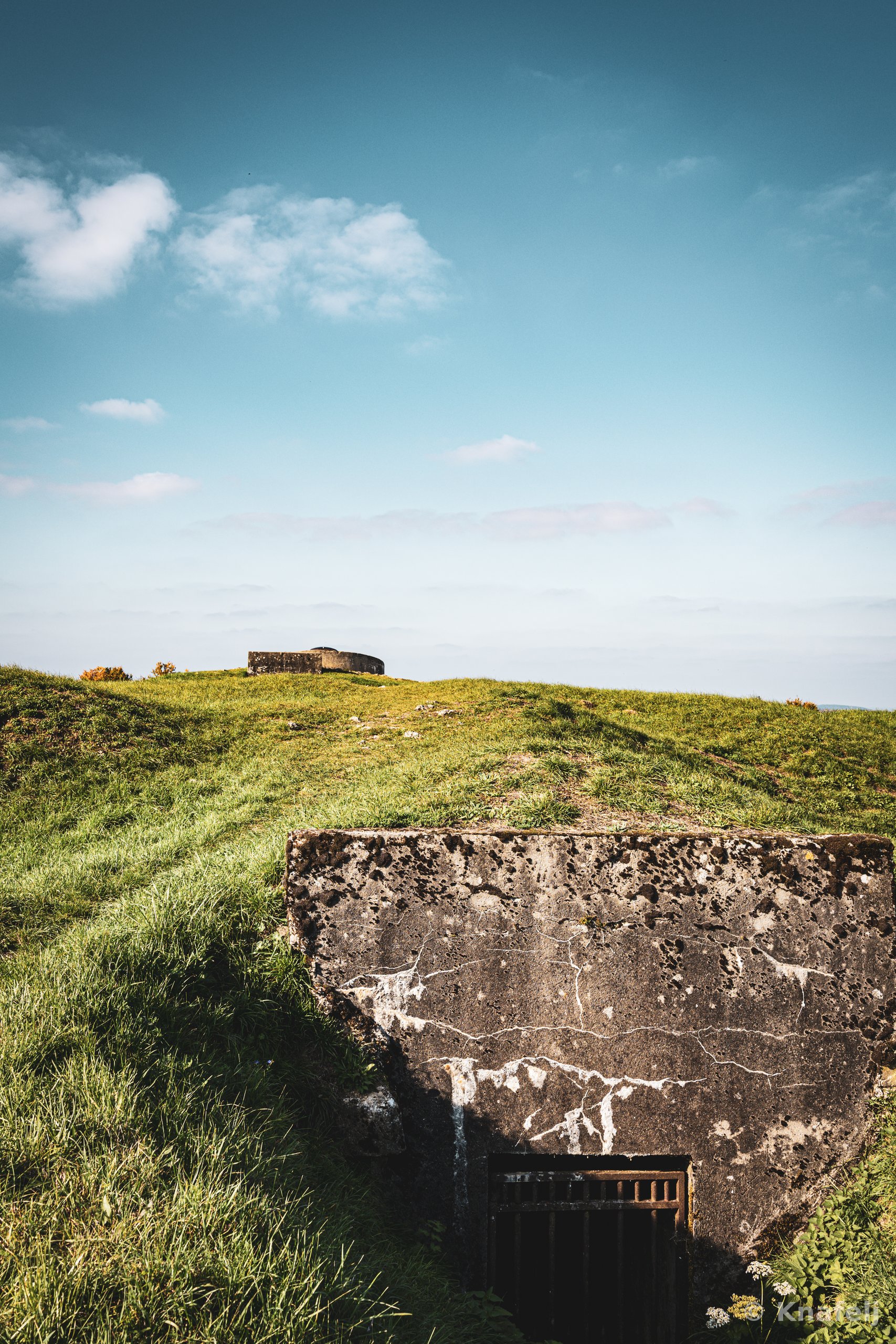 A cracked concrete military bunker built into a grassy hill with a dark doorway, set against a blue sky with scattered clouds, and a second concrete structure on the horizon.