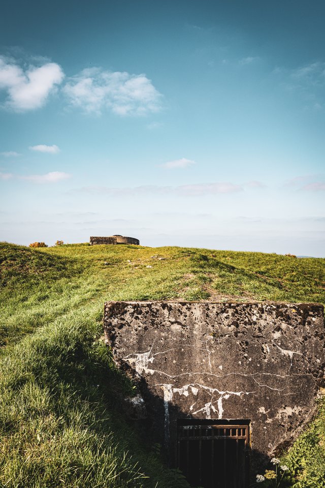 A cracked concrete military bunker built into a grassy hill with a dark doorway, set against a blue sky with scattered clouds, and a second concrete structure on the horizon.