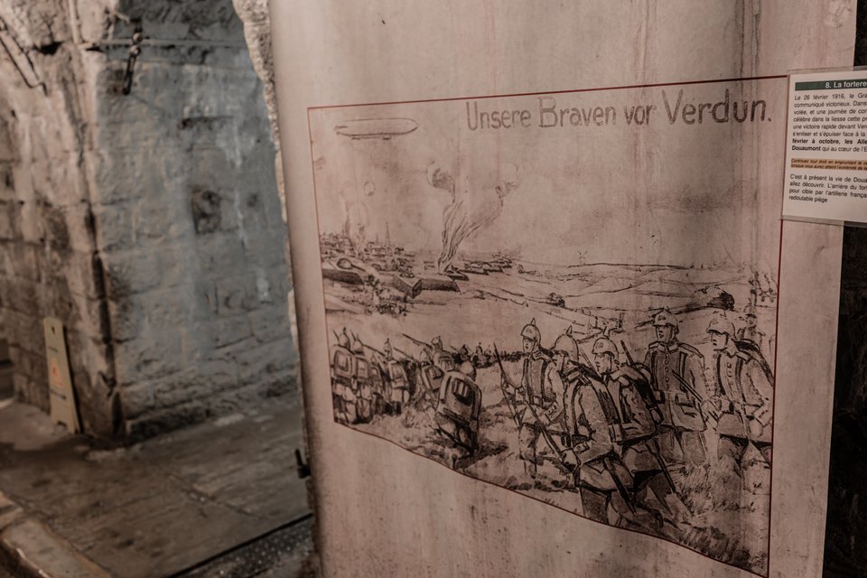 A sepia-toned exhibit panel titled 'Unsere Braven vor Verdun' showing a drawing of soldiers in spiked helmets marching through a battlefield with a zeppelin overhead, mounted on a wall inside a stone museum interior.