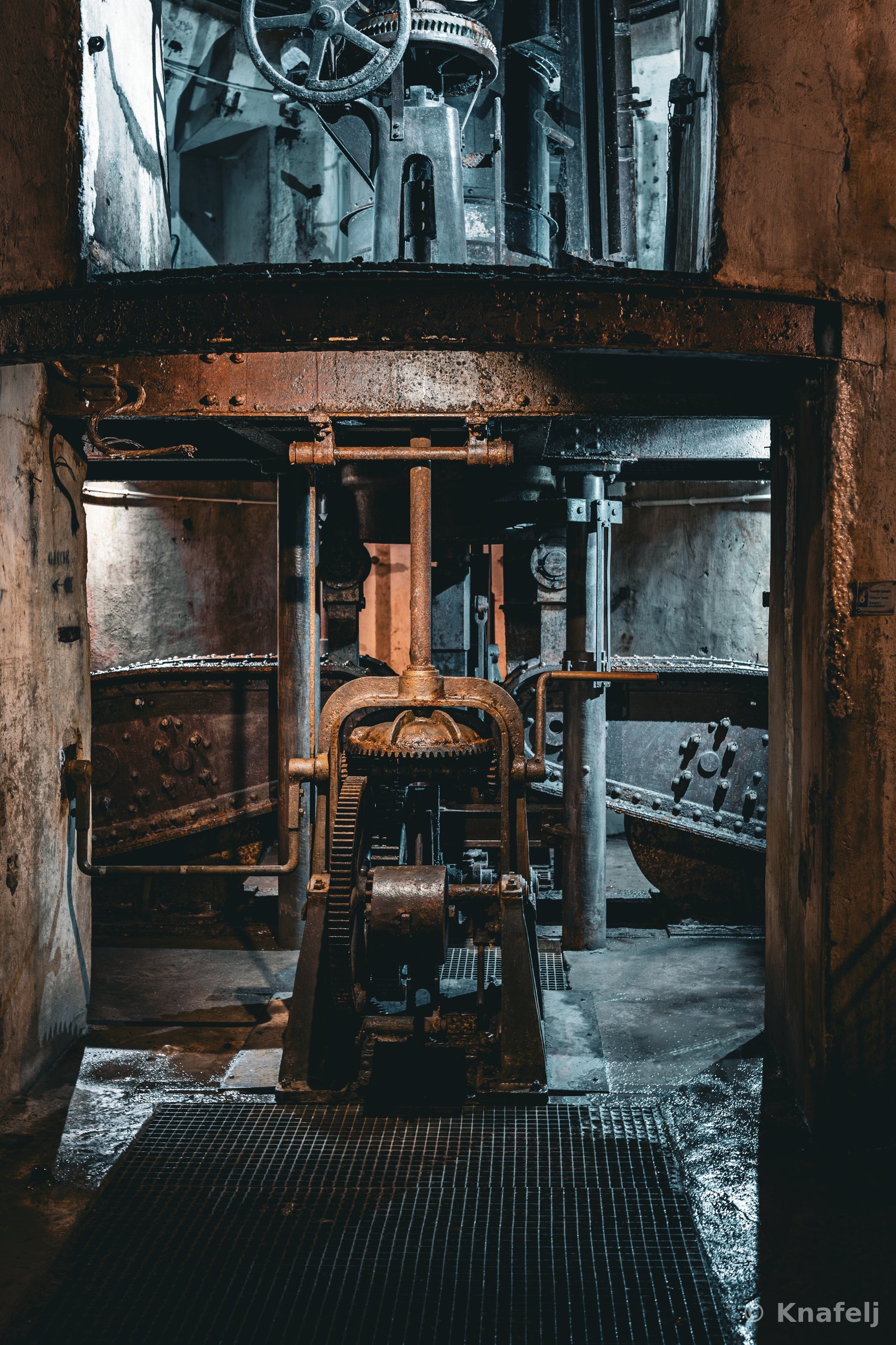 Large rusted industrial machinery with gears, chains, and pipes inside a dark underground fortress chamber, featuring a metal grating floor and weathered concrete walls.