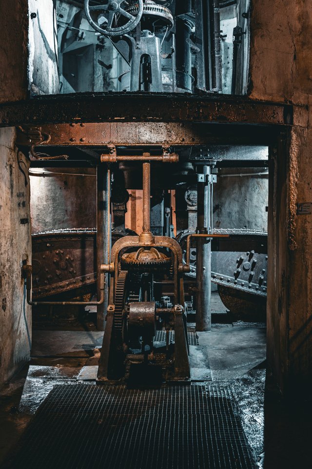 Large rusted industrial machinery with gears, chains, and pipes inside a dark underground fortress chamber, featuring a metal grating floor and weathered concrete walls.