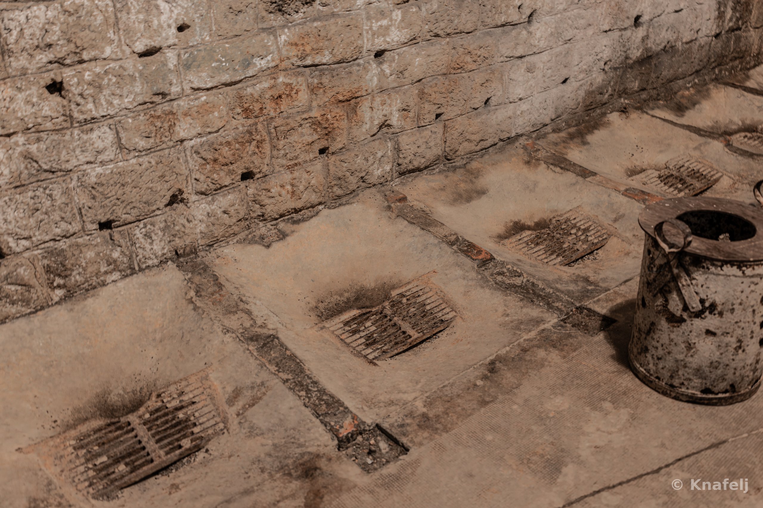 Old stone floor with several rusted metal drain grates and a weathered metal bucket beside a rough brick wall in dim warm light.