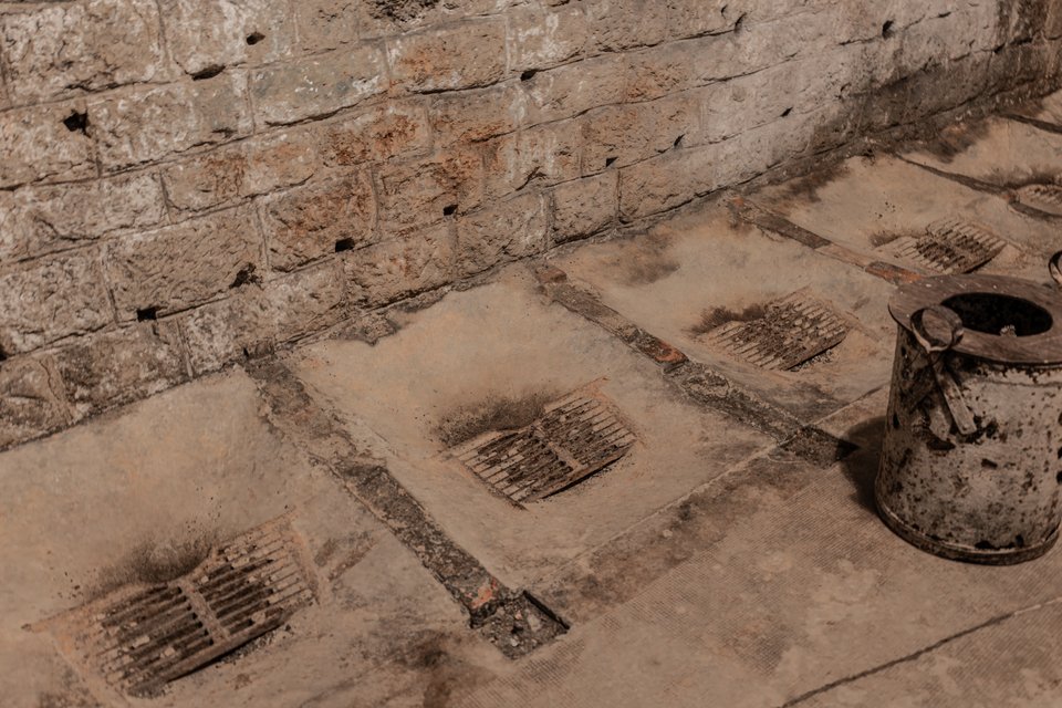 Old stone floor with several rusted metal drain grates and a weathered metal bucket beside a rough brick wall in dim warm light.