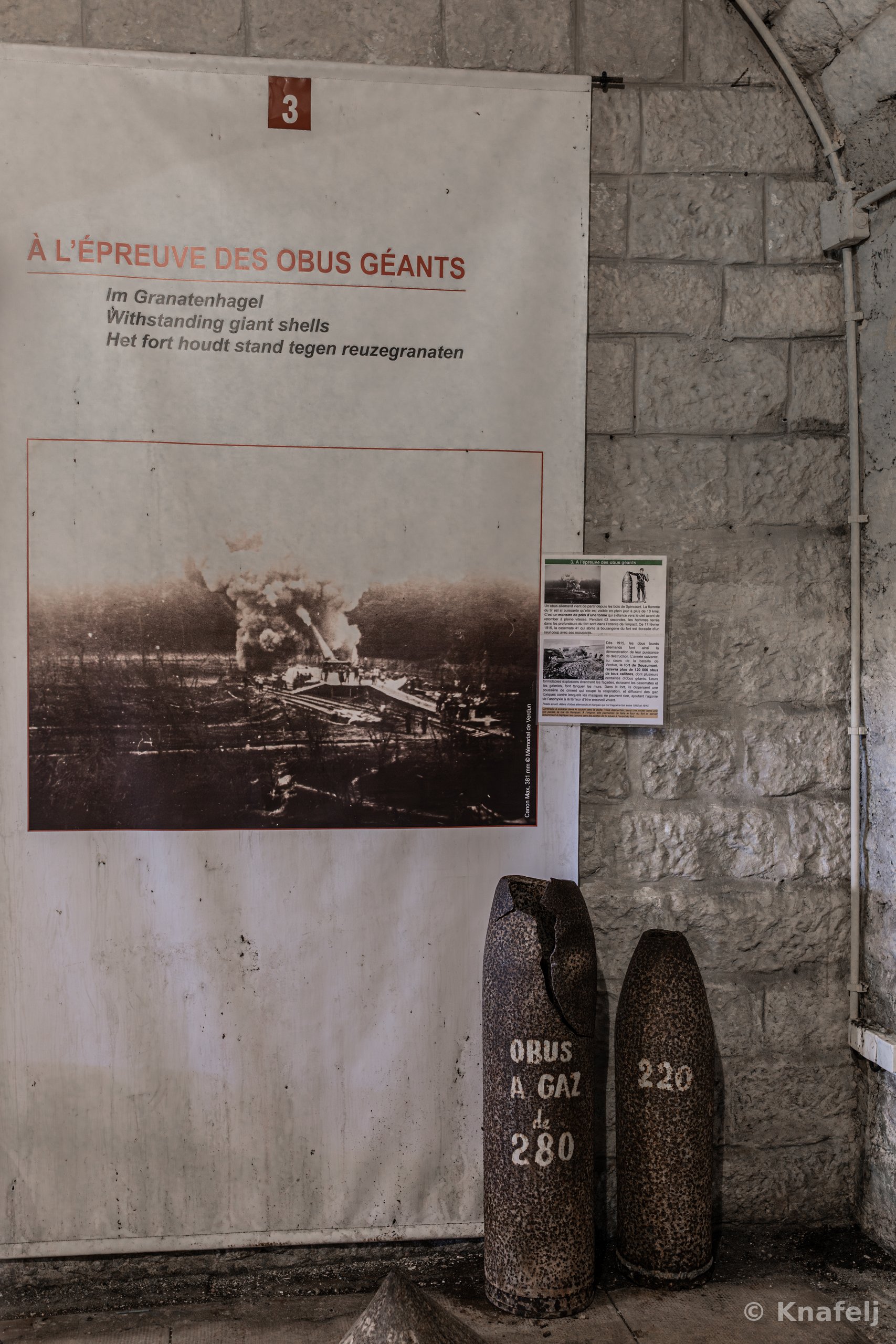 A faded exhibit poster numbered 3 with multilingual text about giant shells hangs on a stone wall, showing a black and white image of an exploding fort. Two large rusted artillery shells labeled 'OBUS A GAZ de 280' and '220' stand on the floor beside it.