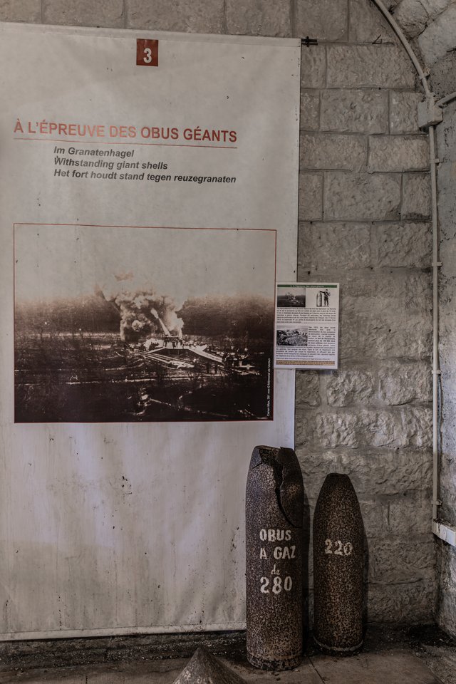 A faded exhibit poster numbered 3 with multilingual text about giant shells hangs on a stone wall, showing a black and white image of an exploding fort. Two large rusted artillery shells labeled 'OBUS A GAZ de 280' and '220' stand on the floor beside it.