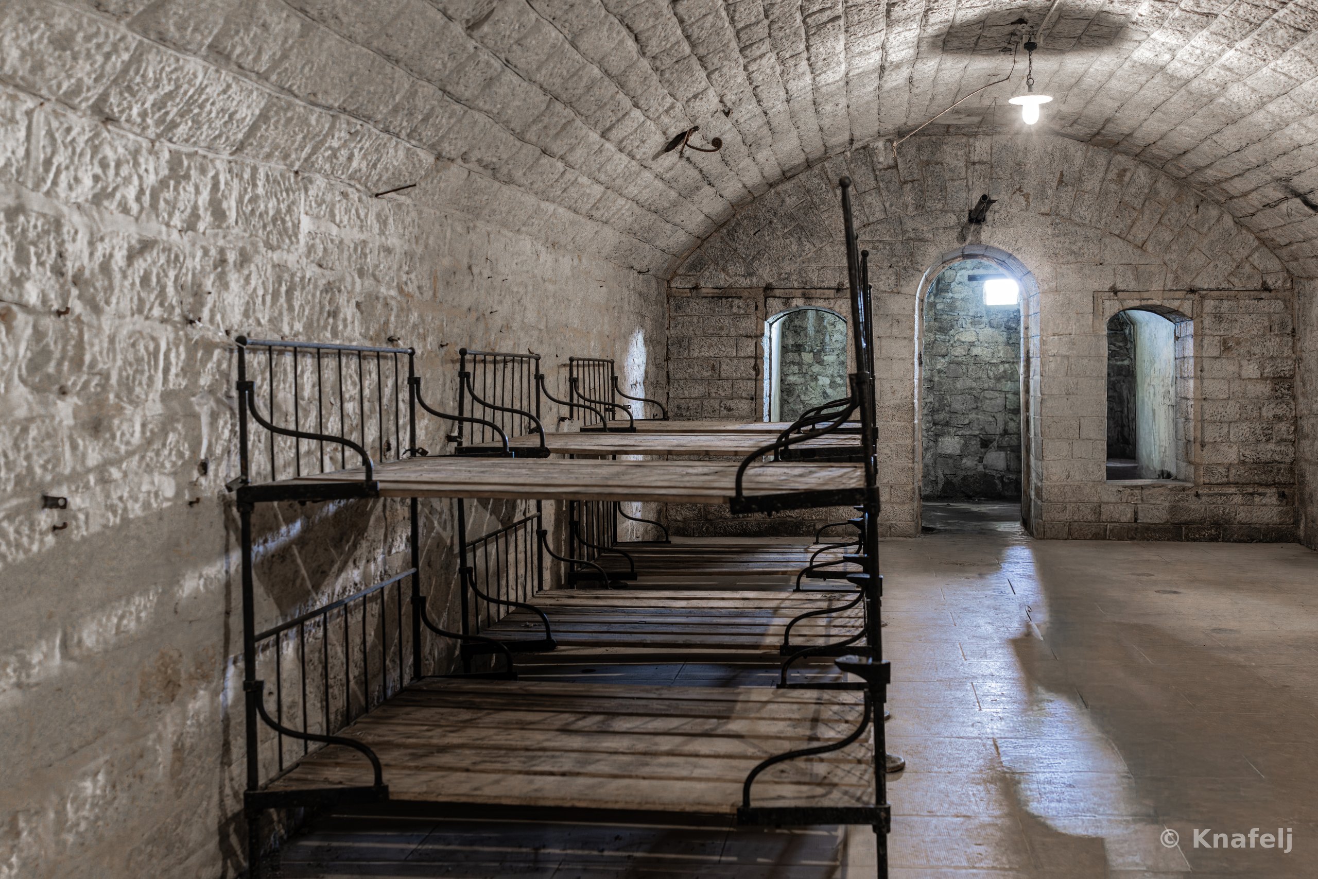 Interior of a vaulted stone room with arched ceiling containing multiple wrought-iron bunk beds with wooden slats, arched doorways and windows in the background, and a single light bulb hanging from the ceiling.