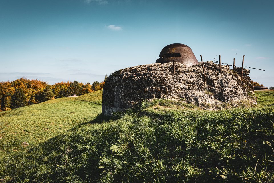 An old concrete military bunker with a rusty steel observation turret on a green grassy hill, surrounded by autumn trees under a blue sky.