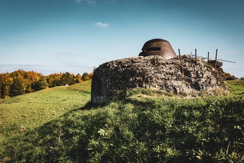 An old concrete military bunker with a rusty steel observation turret on a green grassy hill, surrounded by autumn trees under a blue sky.