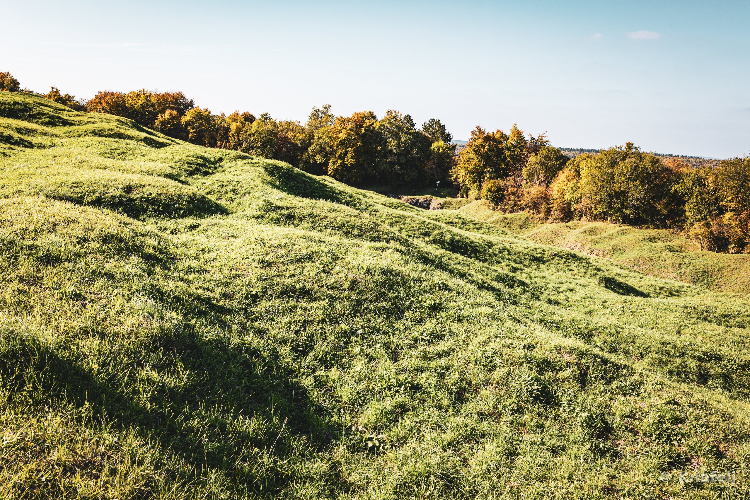 Green rolling hills with uneven grassy mounds and shadows, lined by trees with green and orange foliage beneath a pale blue sky.