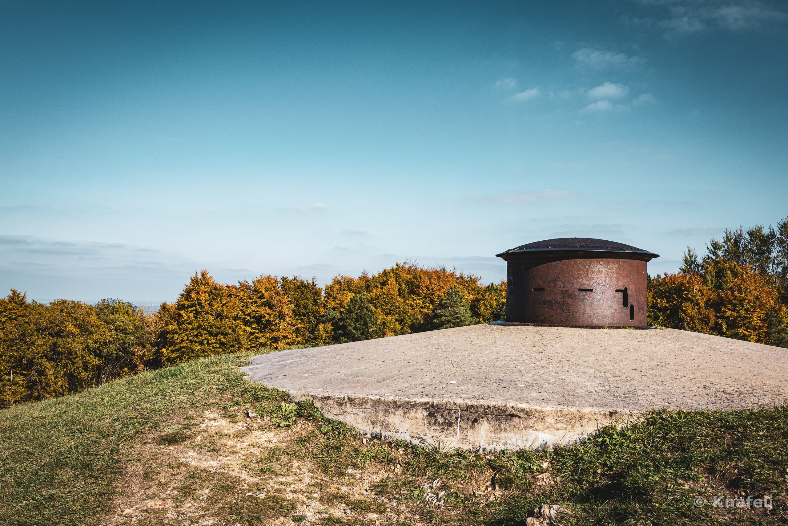 A rusted metal armored cupola mounted on a circular concrete bunker platform atop a grassy hill, with autumn-colored trees in the background under a clear blue sky.