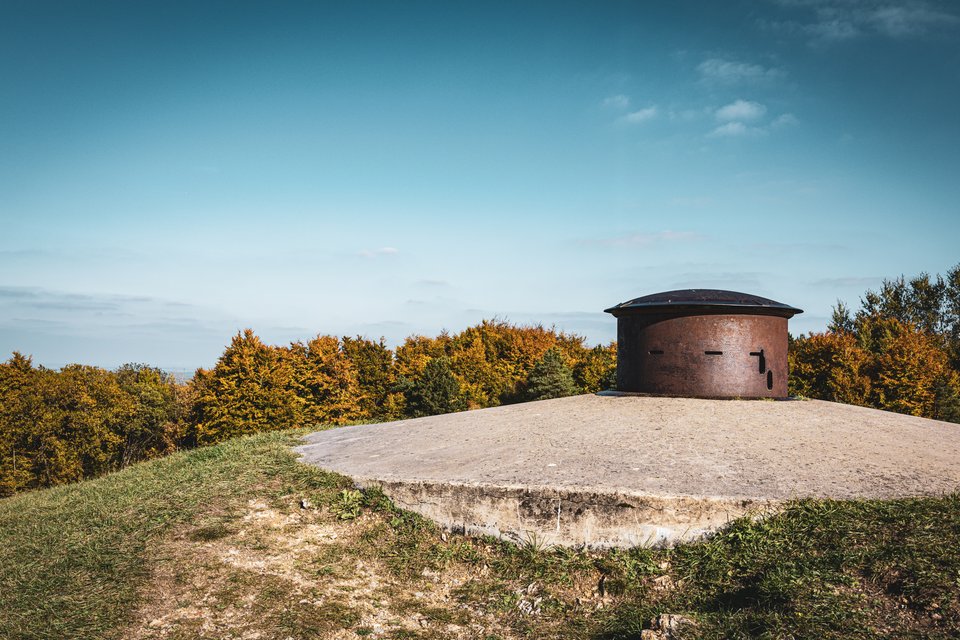 A rusted metal armored cupola mounted on a circular concrete bunker platform atop a grassy hill, with autumn-colored trees in the background under a clear blue sky.