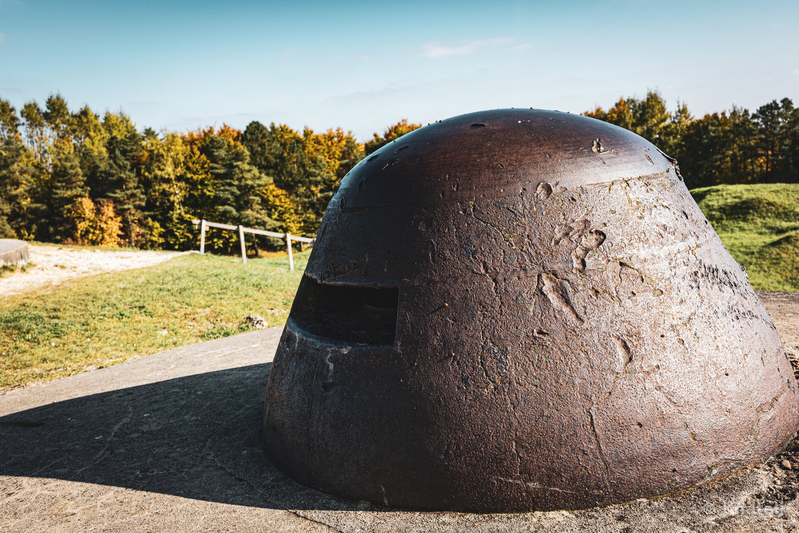 A large rusted steel military bunker dome with an observation slit, set in a concrete platform on a grassy field with trees in the background under a blue sky.